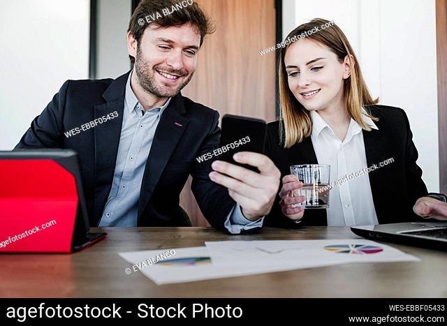 Smiling business colleagues sharing smart phone sitting at desk in coworking office