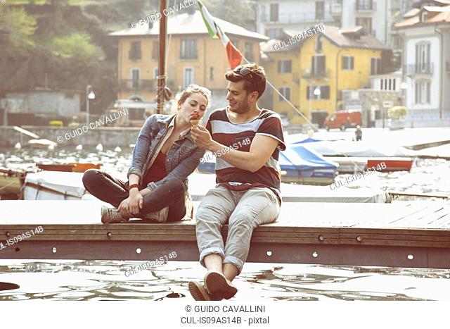 Couple sitting on pier sharing ice cream cone at lake Mergozzo, Verbania, Piemonte, Italy