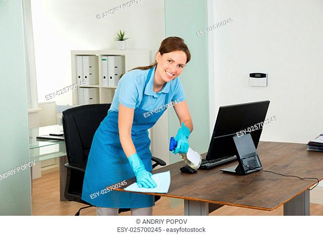 Young Happy Female Janitor Cleaning Wooden Desk With Rag In Office