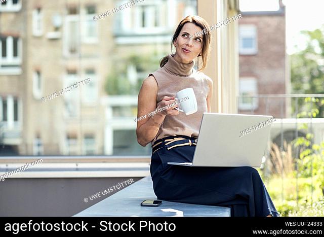 Thoughtful businesswoman sitting with laptop and coffee cup on retaining wall