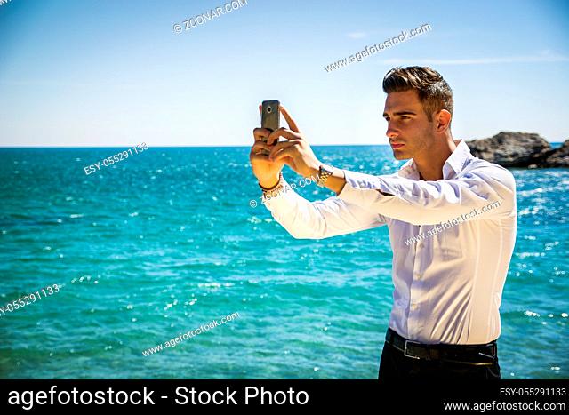 Side View of a Elegant Young Man Taking Selfie Photos at the Beach Standing, Under the Heat of the Sun