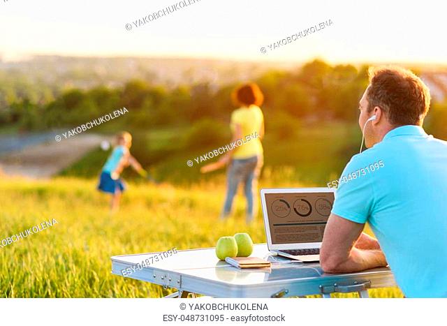 Joining work and leisure. Rear view shot of adult man using laptop and headphones with his wife and daughter playing badminton in background