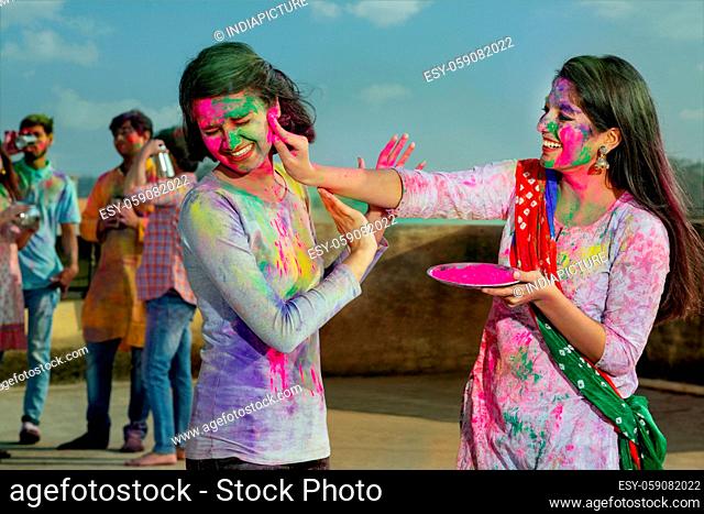 A YOUNG WOMAN PUTTING GULAL ON ANOTHER WOMAN WITH FRIENDS IN BACKGROUND