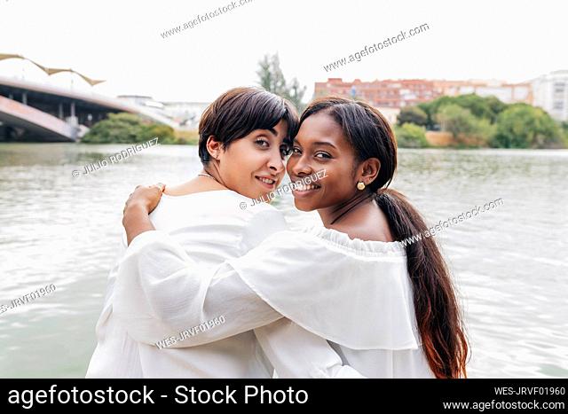 Smiling lesbian couple looking over shoulders at lake