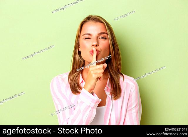 Close-up of blond flirty woman in pink shirt, hushing at camera, share a secret, standing against green background