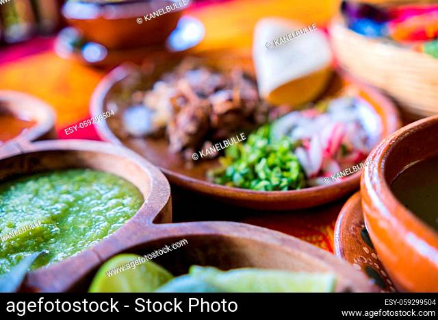 Close-up of brown bowls of lime slices, green sauce, and clay plate of chopped lamb meat on table. Tasty meal and condiments above nice orange and red...
