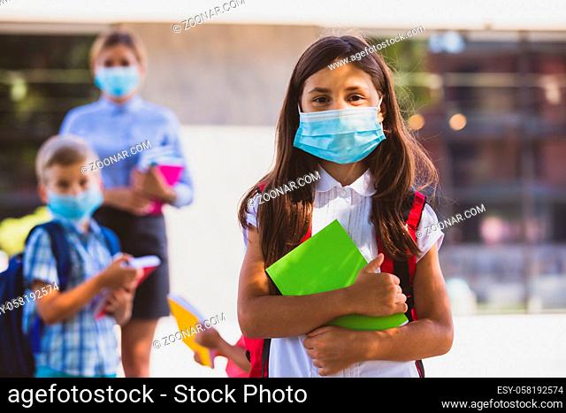 The little schoolgirl with medical mask stands in front of the school. She is standing with backpack and book on the background of a teacher and classmates