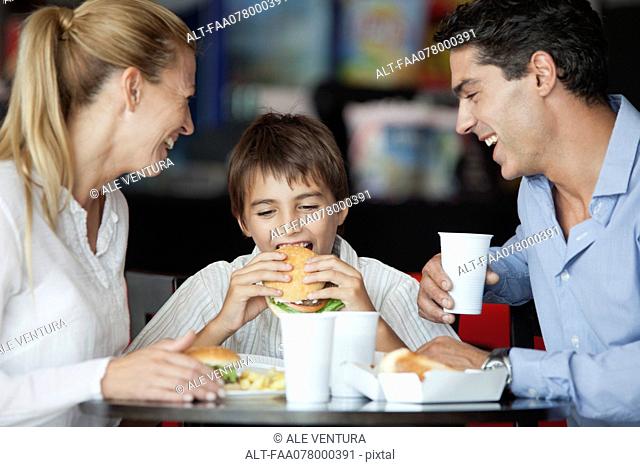 Boy eating hamburger in fast food restaurant with his parents