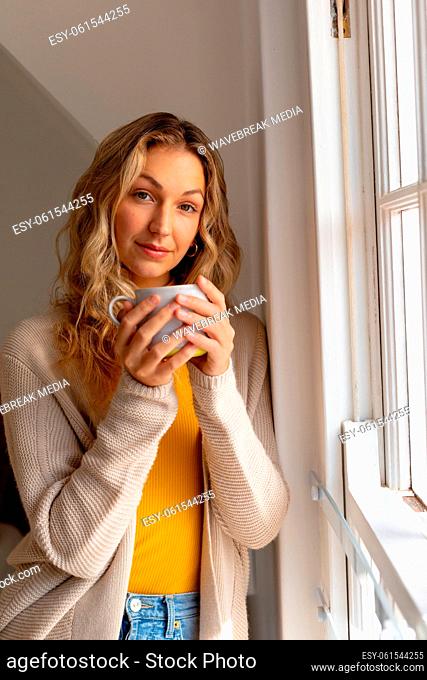 Portrait of happy caucasian woman holding mug, standing next to window and smiling
