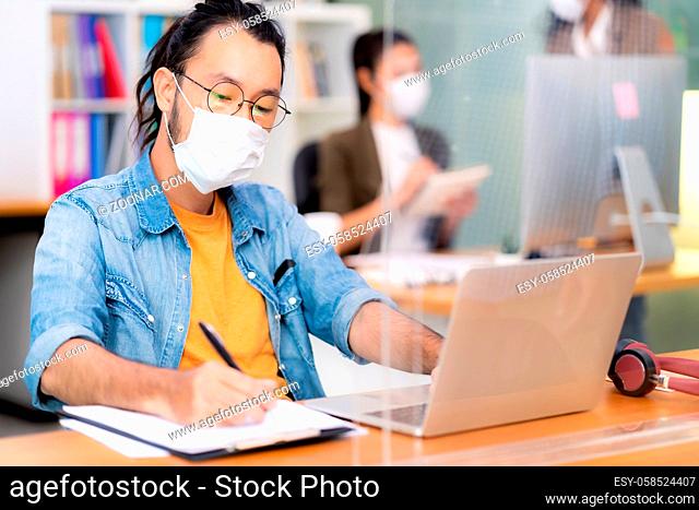 Asian office employee businessman wear protective face mask work in new normal office with interracial colleague in background as social distance practice...