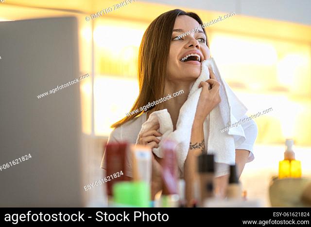 Great idea. Young optimistic woman touching face with towel looking admiringly aside standing near table with laptop and cosmetics
