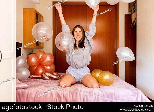 Cheerful woman playing with balloons while kneeling on bed at home