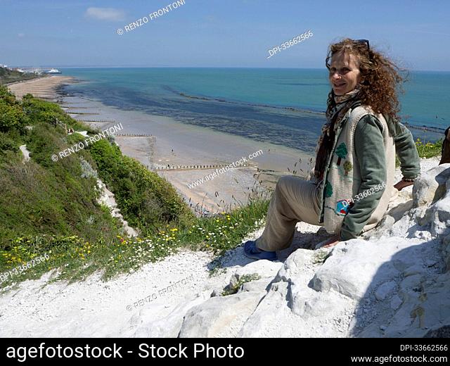Woman sits on white rocks on a ridge above the beach and coastline looking back at the camera; Eastbourne, East Sussex, England