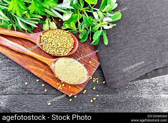 Fenugreek seeds and ground spice in two spoons on brown plate with herbs, a napkin on wooden board background from above