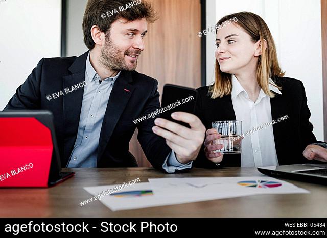 Businesswoman looking at colleague with smart phone discussing sitting at desk in office