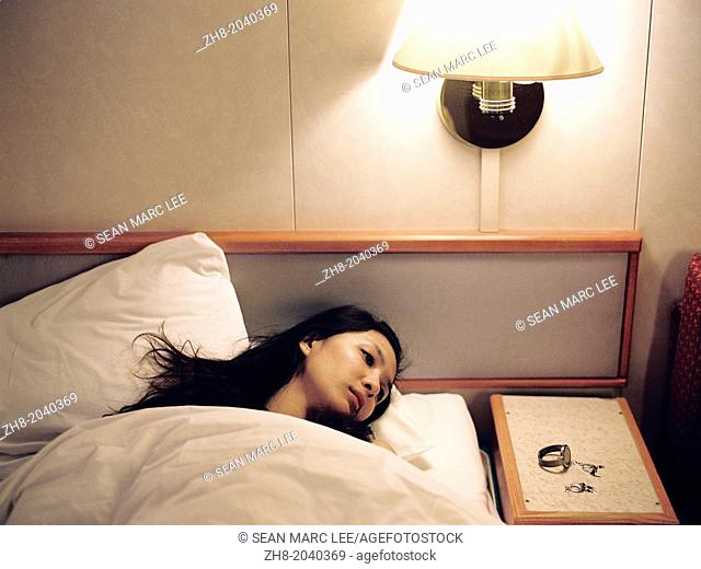 A portrait of a woman lying in a hotel bed with her watch and earrings on the bedside table