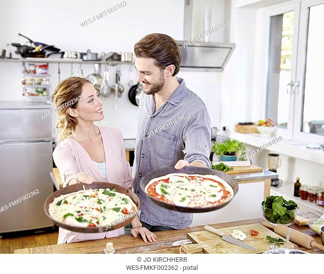 Couple holding baking pans with raw pizzas in kitchen
