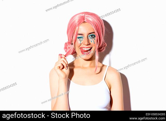 Portrait of sassy attractive girl in pink wig, wearing outfit for halloween party, smiling and winking at camera, standing over white background