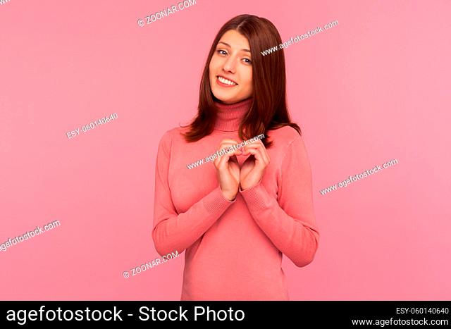 Cute pretty brunette woman in pink sweater showing heart gesture with hands near her chest, symbol of love and charity. Indoor studio shot isolated on pink...