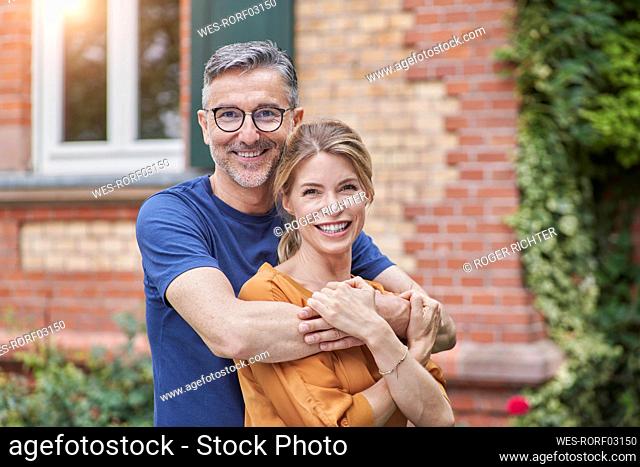 Smiling man embracing woman from behind in front of house