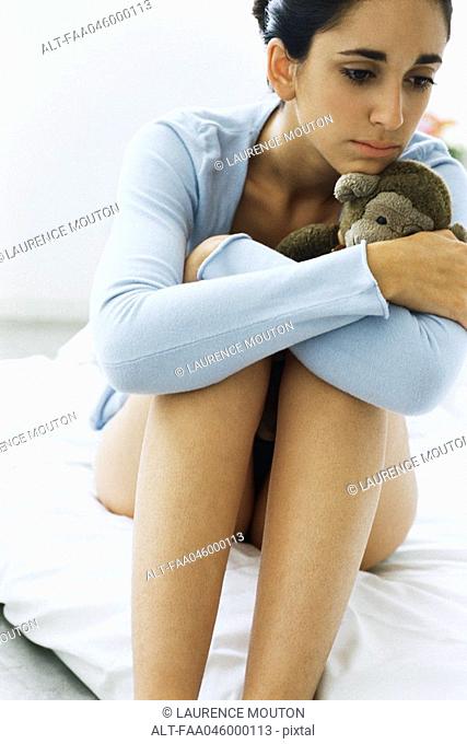Young woman hugging knees, holding stuffed monkey, despondently looking away