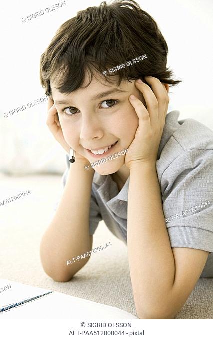 Boy lying on the ground with notebook, holding head, smiling at camera
