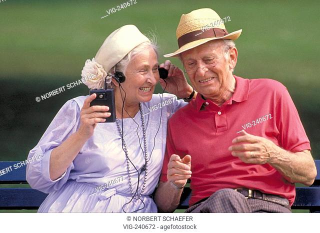 portrait, senior couple wearing hats is sitting on a bench listening amused to music from a walkman using the earphones together  - 0, GERMANY, 24/09/2002