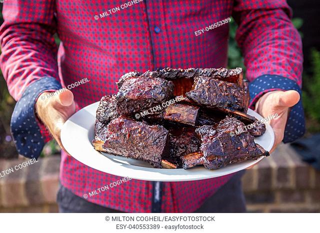 Man carrying a large plate of cooked beef short ribs from the barbecue