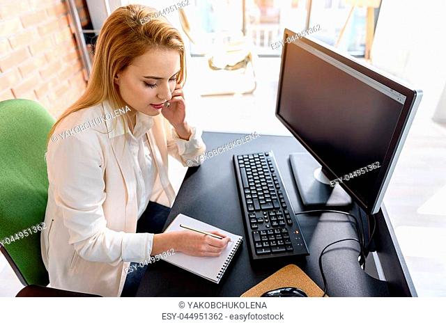 Daily routine. Top view of charming business woman sitting in office and talking by phone while making some notes. She is expressing calm and confidence