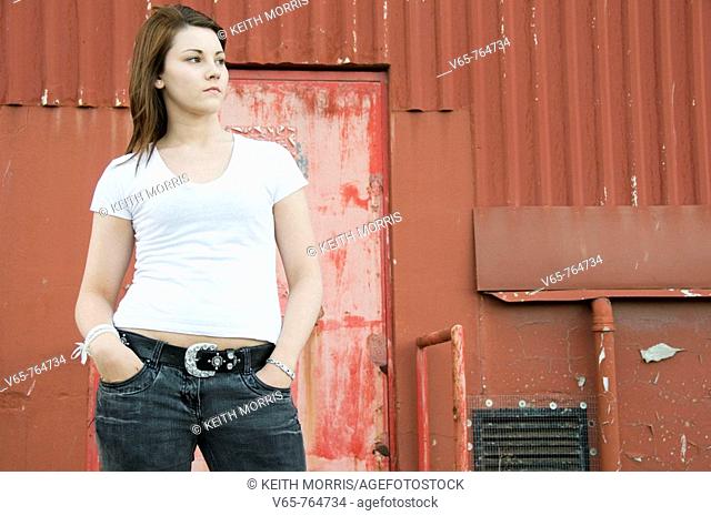 Teenage girl wearing white t-shirt standing in front of red corrugated iron building, looking moody