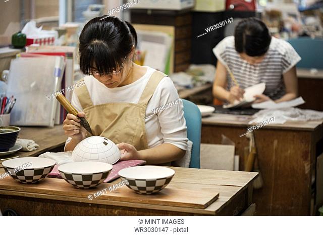 Woman working in a Japanese porcelain workshop, painting geometric pattern onto white bowls with paintbrush