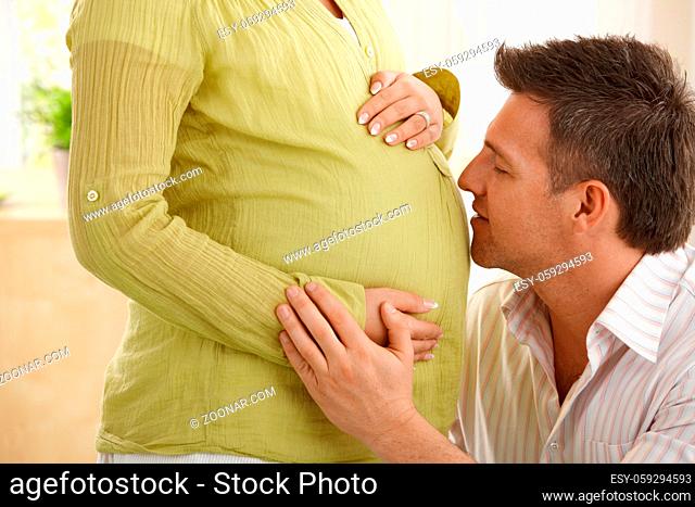 Man talking to baby in expecting wife's tummy, stroking wife's hand in sitting room