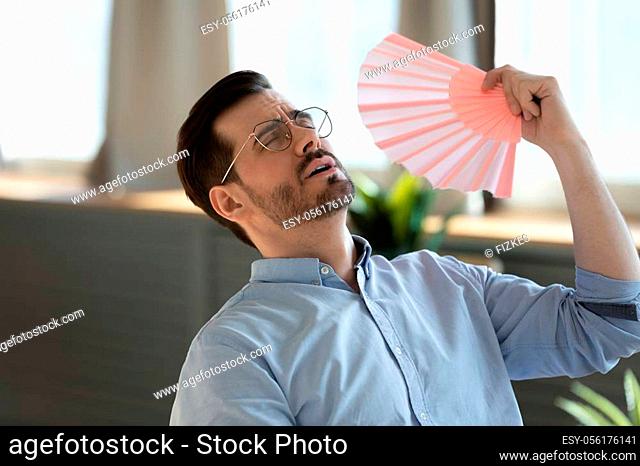 Overheated man in glasses waving paper fan close up, feeling unwell, exhausted tired young male with closed eyes breathing fresh air, suffering from heating