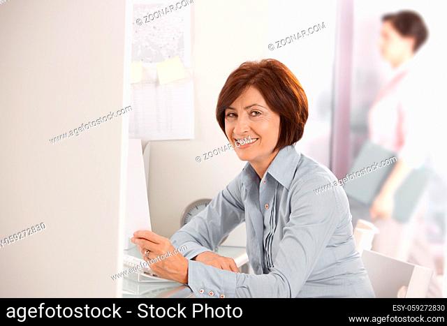Portrait of mature office worker smiling at camera, holding document, colleague in background