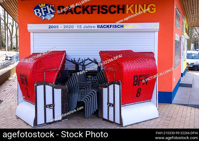 22 March 2021, Mecklenburg-Western Pomerania, Koserow: Beach chairs and terrace chairs stand in front of the closed sales stand for baked fish on the beach near...