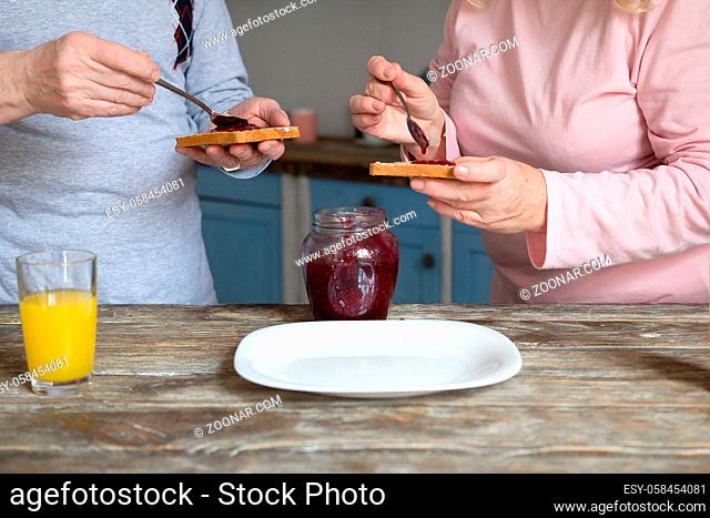 Aged couple making jam with toasts for breakfast. Two senior people prepaing morning meal with jam toasts and orange juice on kitchen wooden table