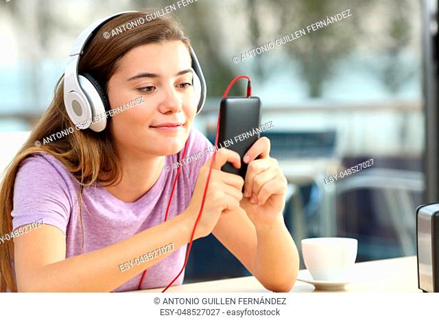 Relaxed teenager wearing headphones is listening to music holding a smart phone in an hotel bar