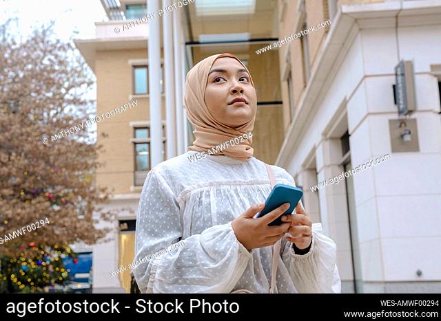 Thoughtful young woman with smart phone standing outside building