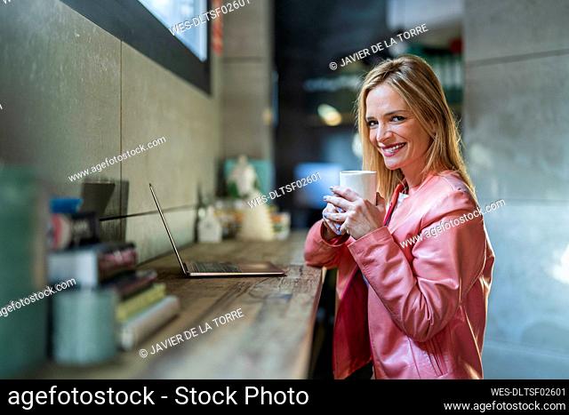 Smiling businesswoman with laptop on desk holding coffee mug at office