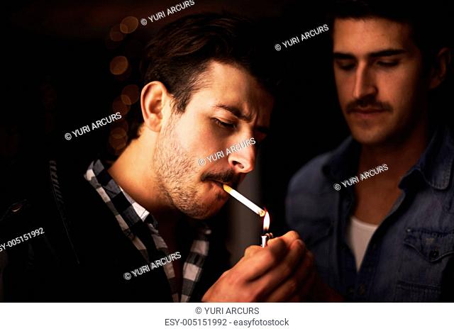 A young guy with a moustache lighting his cigarette as a friend looks on