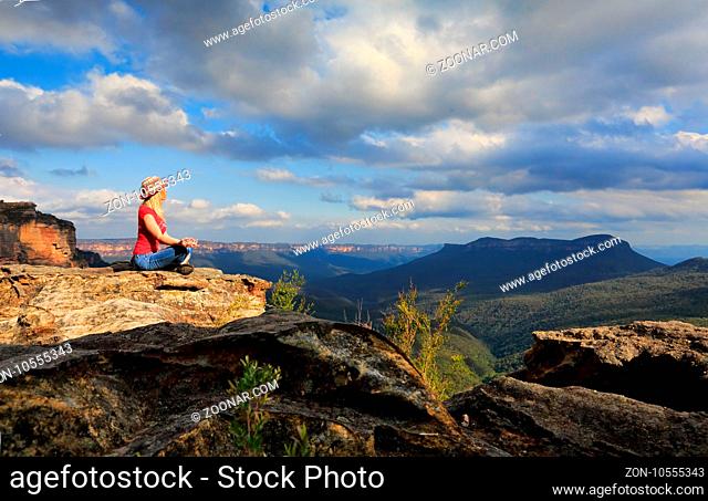 A woman sits peacefully and meditates in a tranquil location on summit of Blue Mountains on a rocky precipice with scenic views