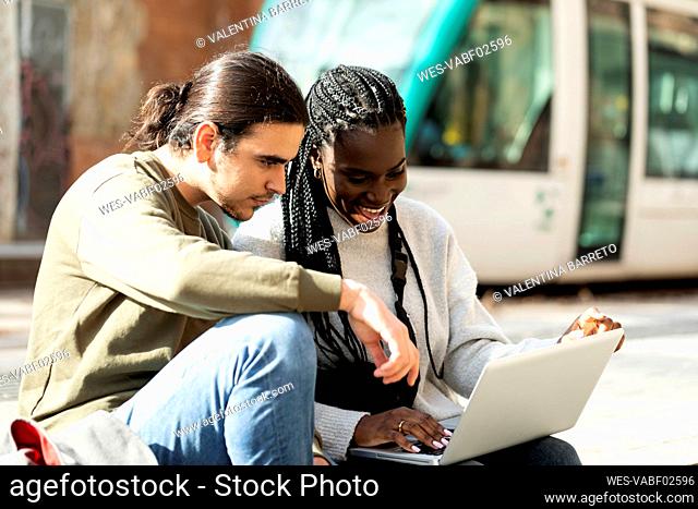 Happy young couple using laptop at tram stop in the city