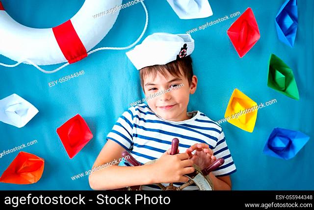seven years old boy playing in sailor hat. Child with a steering wheel and paper ships on blue background