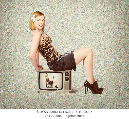 Creative image of a young blond retro pinup woman sitting on a 1950s vintage tv set while tuning to a less static frequency. Television show actress
