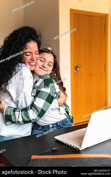 Smiling mother embracing daughter in front of laptop at home