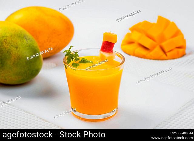 A glass filled with mango juice in the foreground and in the background two whole mangoes on one side and a half mango on the other side
