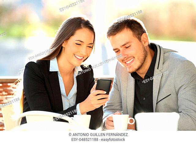 Two happy executives watching on line content in a mobile phone sitting in a restaurant terrace