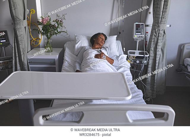 Front view of mature mixed race female patient sleeping in bed with one hand on her stomach in the ward at hospital. Flowers