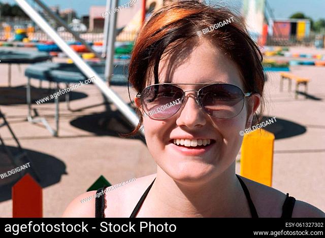 Young woman with black orange hair, wearing sunglasses, smiling, looking at camera while standing in front of a trampoline park in majorca, close-up