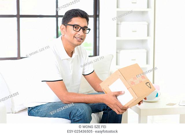 Courier delivery concept. Indian guy received an express parcel and checking the box at home. Handsome male portrait. Asian man sitting on sofa indoor